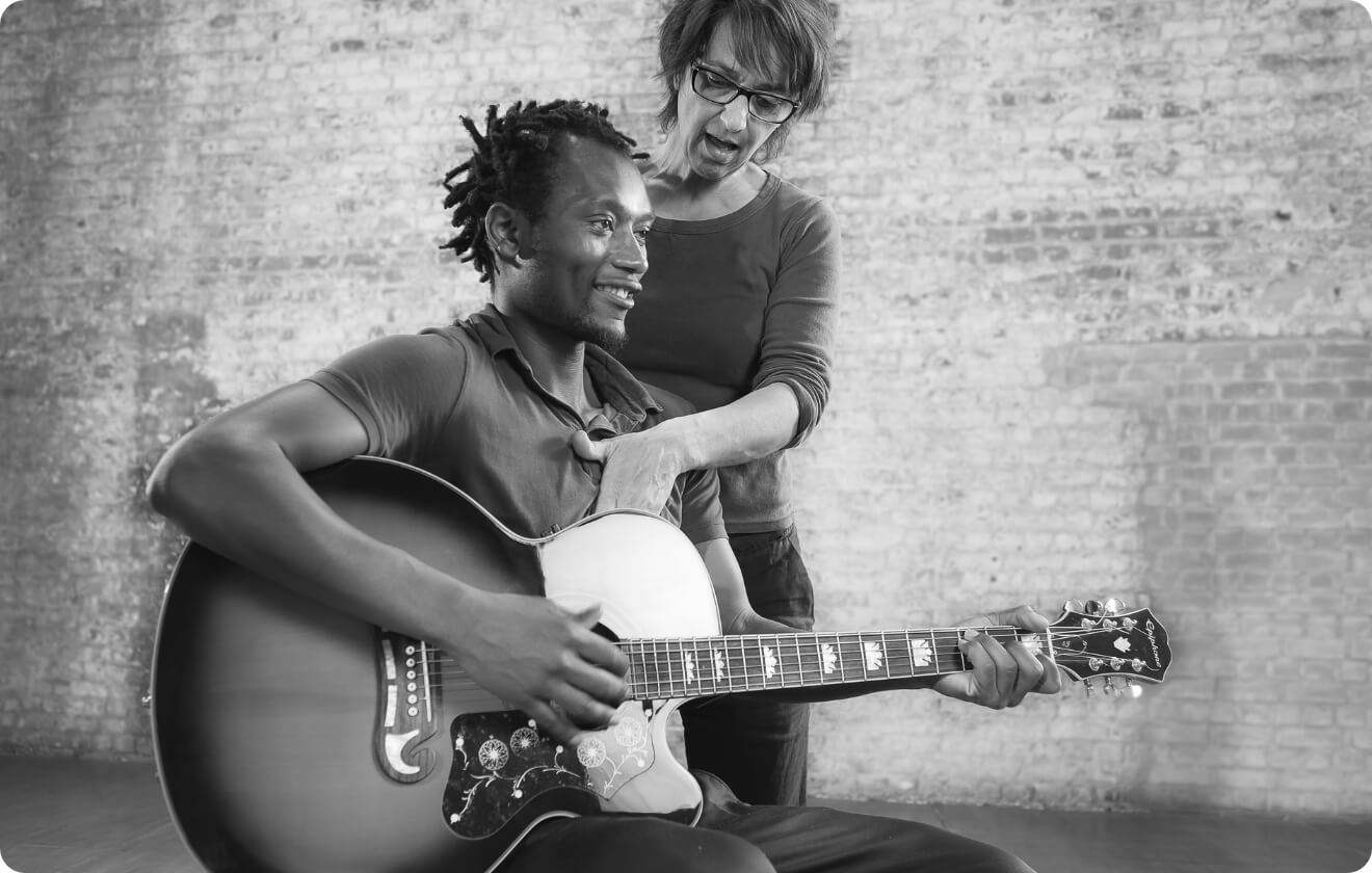 Fotograph: Robert Golden Ein Gitarrist spielt auf seiner Gitarre, während eine Feldenkrais-Lehrerin ihn sanft am Oberkörper unterstützt. Die Szene zeigt, wie Körperbewusstsein und Bewegungsschulung in die musikalische Praxis integriert werden.