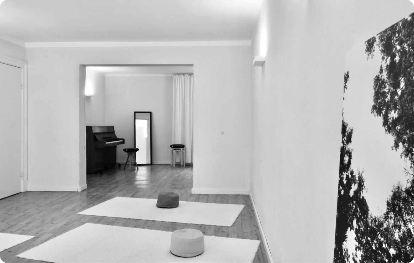 Heller Übungsraum mit Matten und Meditationskissen auf Holzboden, im Hintergrund Klavier und Spiegel.